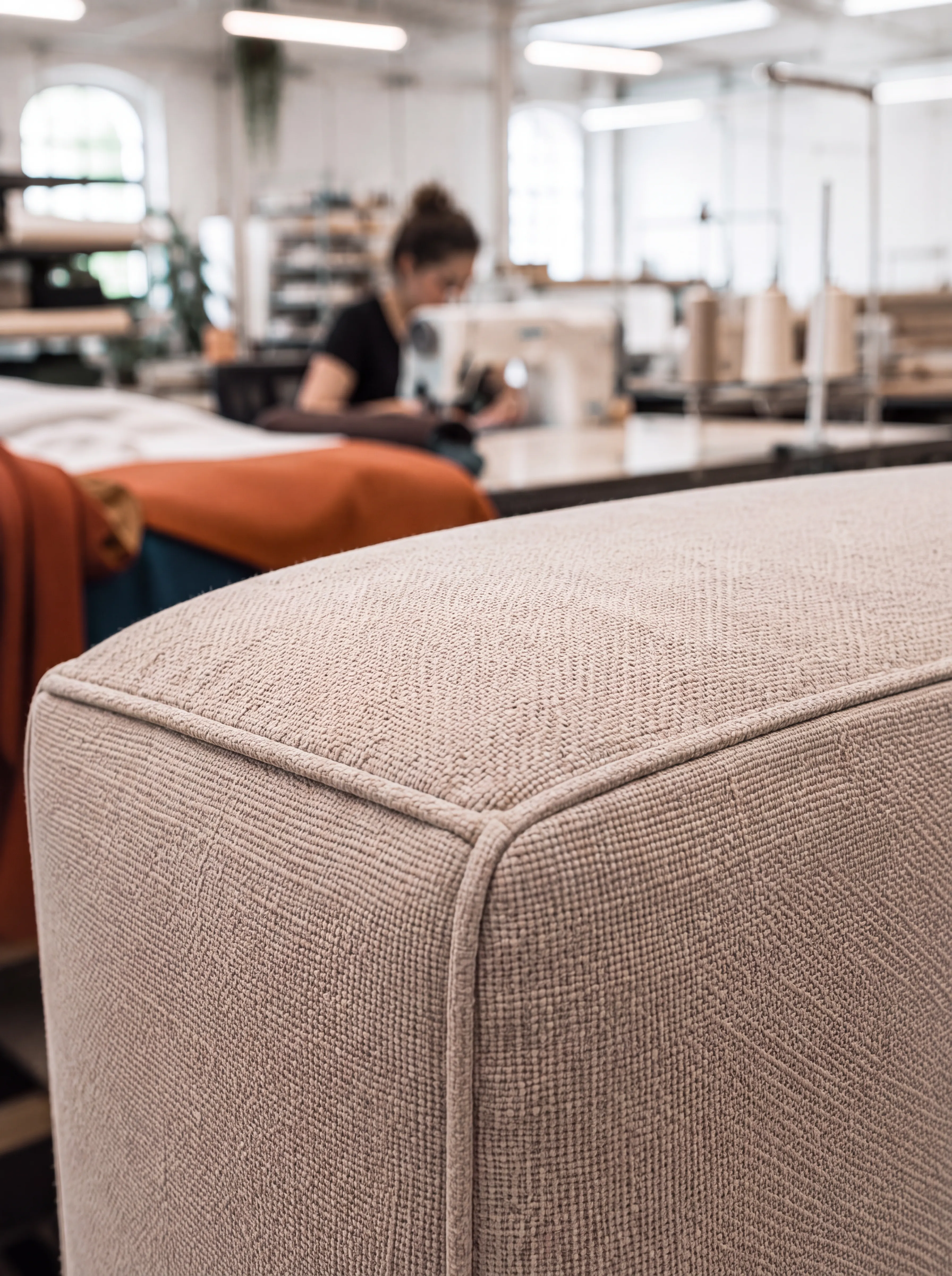 Close-up of a BELANDA sofa arm in oatmeal upholstery, showing the piped seam and cushion edge, with a seamstress at work behind it